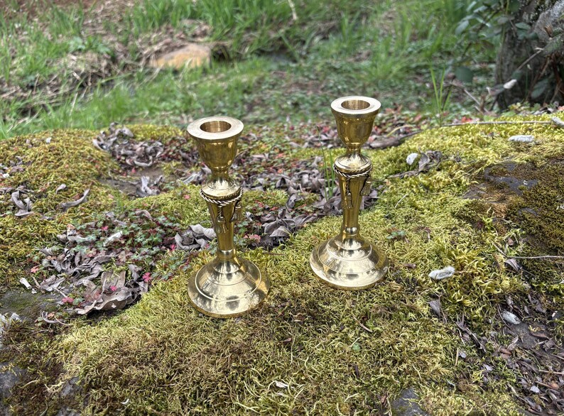 May include: Two antique brass candle holders. The candle holders have a detailed design with a round base, a slender stem, and a cup-shaped top. The candle holders are placed on a bed of moss.