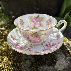 K&ouml;nnte beinhalten: Ein wei&szlig;es Tee-Set mit rosa und gr&uuml;nen Blumenmustern. Tasse und Untertasse haben einen Goldrand. Die Tasse steht auf der Untertasse. Das Set steht auf einem moosbewachsenen Stein.