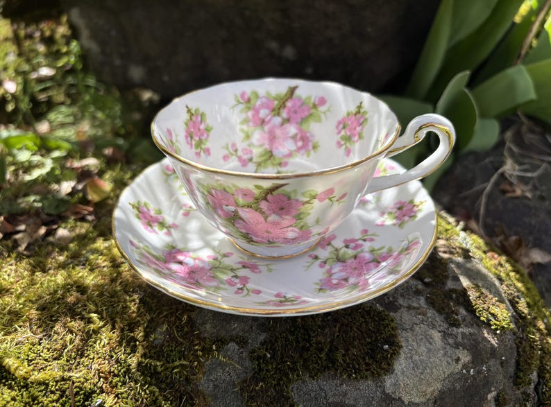 K&ouml;nnte beinhalten: Ein wei&szlig;es Tee-Set mit rosa und gr&uuml;nen Blumenmustern. Tasse und Untertasse haben einen Goldrand. Die Tasse steht auf der Untertasse. Das Set steht auf einem moosbewachsenen Stein.