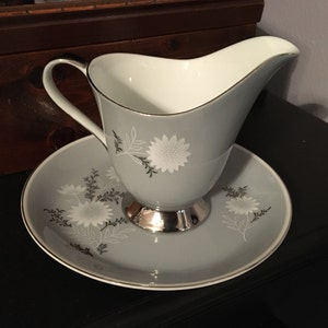 May include: A grey ceramic creamer with a silver base and white floral design. The creamer is on a matching saucer.