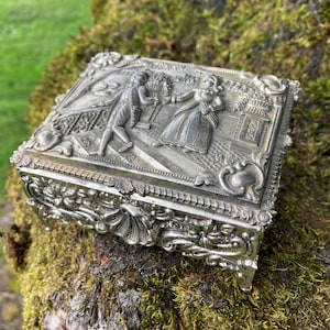 May include: A silver metal trinket box with an ornate design. The top of the box features a raised relief of two figures in a garden setting.