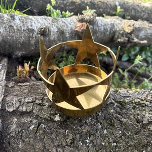 Puede incluir: Un portavelas de metal dorado con dise&ntilde;o de estrella. El portavelas tiene una base circular y una parte superior en forma de corona con tres estrellas grandes y un anillo de estrellas m&aacute;s peque&ntilde;as. El portavelas est&aacute; sobre un tronco desgastado.