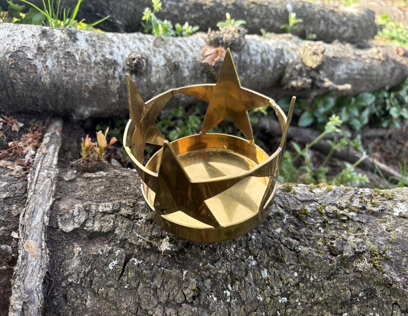 Puede incluir: Un portavelas de metal dorado con dise&ntilde;o de estrella. El portavelas tiene una base circular y una parte superior en forma de corona con tres estrellas grandes y un anillo de estrellas m&aacute;s peque&ntilde;as. El portavelas est&aacute; sobre un tronco desgastado.