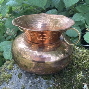Puede incluir: Jarra de cobre martillado con cuerpo redondo y borde acampanado. Tiene un asa de latón y está sobre una roca cubierta de musgo. El fondo presenta follaje verde.