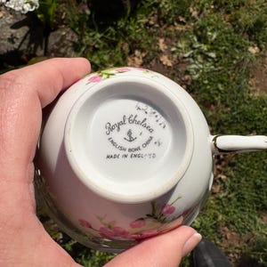 K&ouml;nnte beinhalten: Eine wei&szlig;e Royal Chelsea English Bone China Teetasse mit rosa Blumenmustern. Auf der Unterseite der Tasse befinden sich der Markenname, ein Anker und die Aufschrift "MADE IN ENGLAND". Der Griff ist wei&szlig; mit goldenen Details.
