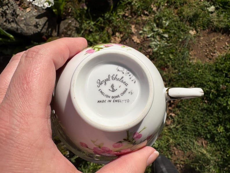 K&ouml;nnte beinhalten: Eine wei&szlig;e Royal Chelsea English Bone China Teetasse mit rosa Blumenmustern. Auf der Unterseite der Tasse befinden sich der Markenname, ein Anker und die Aufschrift "MADE IN ENGLAND". Der Griff ist wei&szlig; mit goldenen Details.