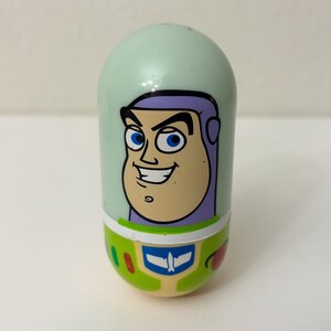 May include: A mint green, oval-shaped toy featuring a cartoon image of Buzz Lightyear from Toy Story. The toy has a smiling face, purple accents, and a green and white design at the base, with a blue and white emblem.