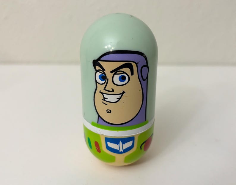 May include: A mint green, oval-shaped toy featuring a cartoon image of Buzz Lightyear from Toy Story. The toy has a smiling face, purple accents, and a green and white design at the base, with a blue and white emblem.