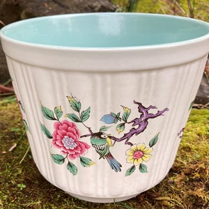 Könnte beinhalten: Weißer Keramiktopf mit hellblauem Innenraum. Der Topf zeigt ein florales Design mit einem Vogel und einem Zweig in den Farben Rosa, Grün, Gelb, Lila und Blau.