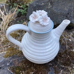 May include: White ceramic teapot with a floral lid. The teapot has a speckled glaze and a blue trim around the lid. The lid is decorated with two white ceramic flowers.