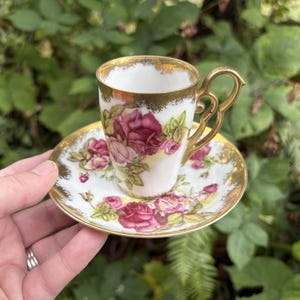 Könnte beinhalten: Ein Mokkatassen-Set aus weißem Porzellan mit einem Blumenmuster aus rosa und dunkelroten Rosen sowie grünen Blättern. Der Rand der Tasse und der Untertasse ist mit einer Goldverzierung versehen, und die Tasse verfügt über einen goldenen Henkel.