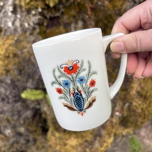 May include: White ceramic mug with a floral design featuring red, blue, and orange flowers. The mug has a curved handle and is held against a natural background. The floral pattern is centered on the mug's front.
