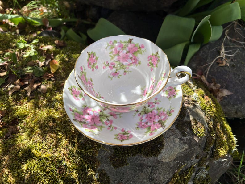 K&ouml;nnte beinhalten: Ein wei&szlig;es Tee-Set mit rosa Blumenmuster und goldenem Rand. Die Tasse steht auf der Untertasse, beide auf einem moosbewachsenen Felsen. Die Tasse hat einen zarten Henkel.