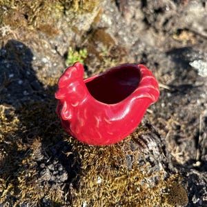 May include: A small, glossy red ceramic figurine shaped like a chicken, featuring a detailed head with a comb and a rounded body with a hollow opening at the top. The surface is smooth and reflective, showcasing a vibrant, solid red color throughout.