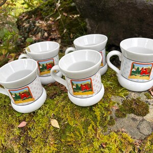 May include: Five white ceramic mugs with handles are arranged on a mossy rock. Each mug features a colorful farm scene with a red barn. The mugs have a patterned band around the middle and a white base.
