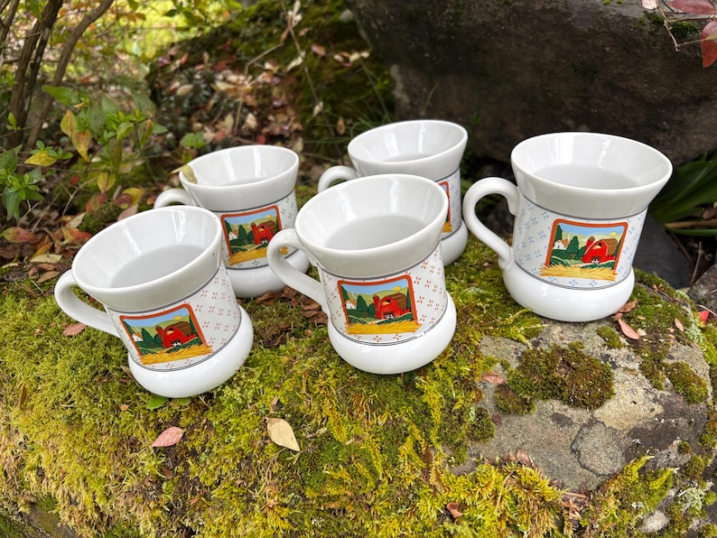 May include: Five white ceramic mugs with handles are arranged on a mossy rock. Each mug features a colorful farm scene with a red barn. The mugs have a patterned band around the middle and a white base.