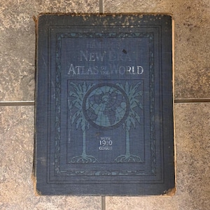 May include: An antique blue hardcover book titled "New Era Atlas of the World" with the year 1910 printed at the bottom. The cover features an illustration of a globe surrounded by trees.