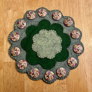 May include: A green and gray felt circle with a floral design. The circle is decorated with 13 wooden buttons with floral patterns.