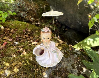 May include: A vintage porcelain figurine of a girl in a pink dress, holding a basket and a small parasol. The figurine has a flower in her hair and is decorated with gold accents. The figurine is sitting on a mossy rock.