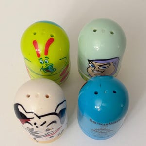 May include: Four colorful, egg-shaped salt and pepper shakers. The shakers are decorated with cartoon character designs in shades of green, blue, white, and yellow. Each shaker has three holes on top for dispensing.