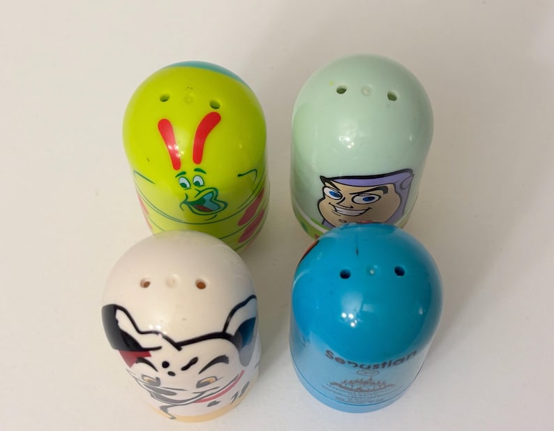 May include: Four colorful, egg-shaped salt and pepper shakers. The shakers are decorated with cartoon character designs in shades of green, blue, white, and yellow. Each shaker has three holes on top for dispensing.