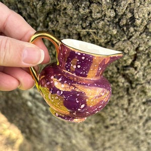 May include: A small, decorative pitcher with a gold handle and rim. The pitcher has a deep purple base with gold and white speckled accents. The interior is white. The pitcher is held against a textured, neutral background.