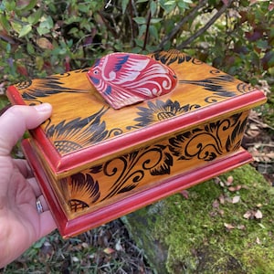 May include: A wooden box with a red and black floral design. The box has a hinged lid with a carved bird design. The box is painted in shades of orange, red, and black.