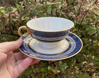 May include: A white teacup and saucer set with a navy blue and gold design. The teacup has a gold handle and a navy blue band with a white speckled pattern. The saucer has a matching navy blue and gold design.
