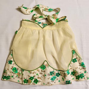 May include: A cream-colored half apron featuring a scalloped hemline with a green and brown floral print. The apron includes a matching tie at the waist and a decorative green trim along the scalloped edges, creating a layered appearance.