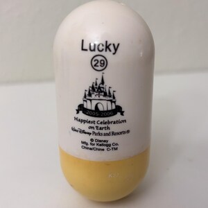 May include: A white and yellow capsule-shaped collectible with the word "Lucky" and the number "29" printed on it. It features a black graphic of a castle and the text "Happiest Celebration on Earth" with "2005-2006" below it.
