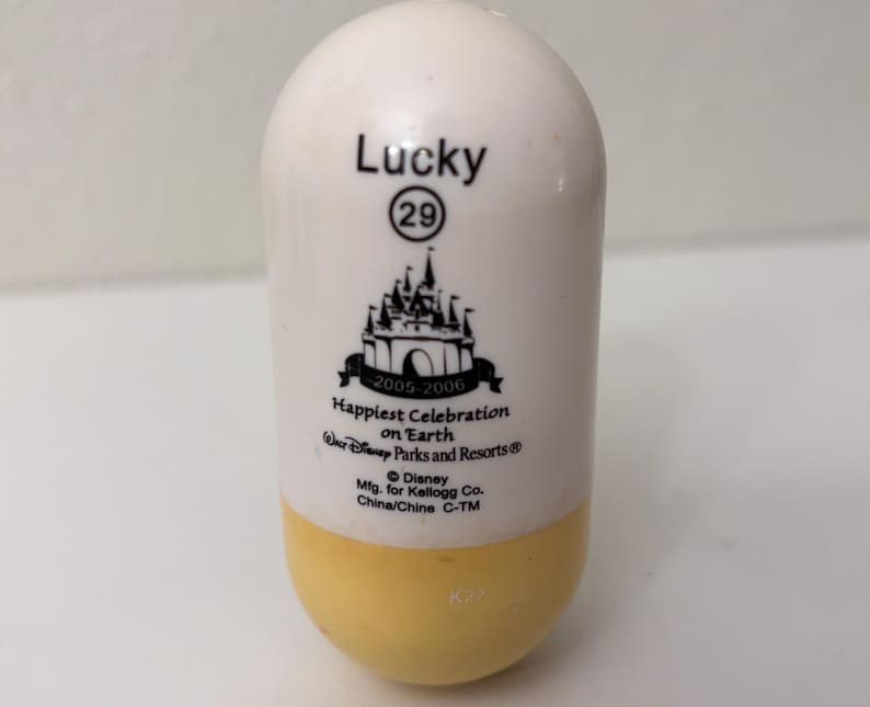May include: A white and yellow capsule-shaped collectible with the word "Lucky" and the number "29" printed on it. It features a black graphic of a castle and the text "Happiest Celebration on Earth" with "2005-2006" below it.