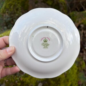 May include: A white ceramic saucer with a scalloped edge. The underside features a central circular design with the text "VIRGINIA STOCK" and "ROYAL STANDARD FINE BONE CHINA ENGLAND". The saucer is held by a hand.