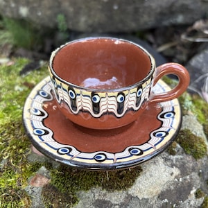 Peut inclure: Un ensemble tasse et soucoupe en céramique marron. La tasse et la soucoupe présentent un motif décoratif de motifs noirs, blancs et bleus. La tasse a une anse et repose sur la soucoupe assortie. L'ensemble est sur une roche moussue.