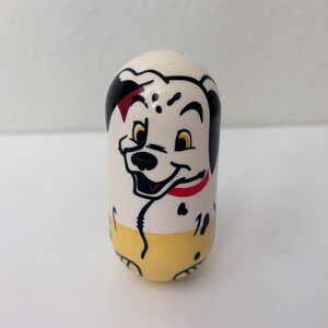 May include: A white, egg-shaped collectible toy featuring a smiling Dalmatian dog face with black spots, a red collar, and yellow pants. The toy has a glossy finish and is set against a plain white background.