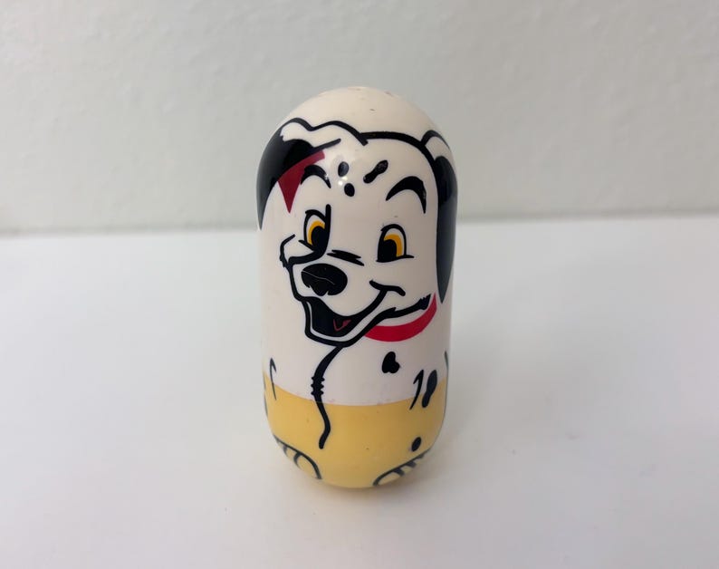 May include: A white, egg-shaped collectible toy featuring a smiling Dalmatian dog face with black spots, a red collar, and yellow pants. The toy has a glossy finish and is set against a plain white background.