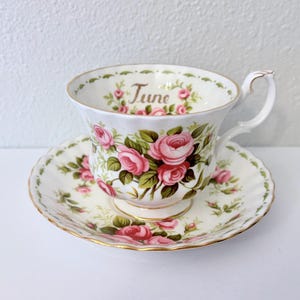 Pode incluir: Um conjunto de chávena e pires brancos com rosas cor-de-rosa e folhas verdes. A chávena tem a palavra "June" impressa. O conjunto tem uma borda dourada.