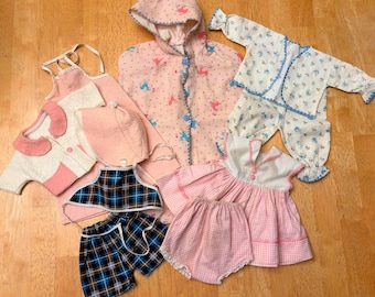 Peut inclure: Une collection de vêtements de poupée comprenant un manteau à capuche rose avec des boutons bleus, un ensemble pyjama à fleurs blanc, une robe rose avec culotte assortie, un tablier rose, un bonnet rose et un haut dos nu à carreaux bleus.