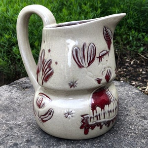 May include: A cream-coloured ceramic jug with a red and white floral design. The jug has a wide mouth and a handle. The design features stylised flowers and leaves.
