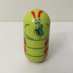 May include: A yellow, capsule-shaped toy with a cartoon caterpillar design. The toy features a surprised face, red accents, and green stripes. The caterpillar's mouth is open, revealing a blue interior.