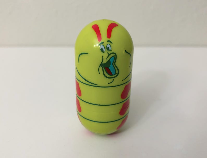 May include: A yellow, capsule-shaped toy with a cartoon caterpillar design. The toy features a surprised face, red accents, and green stripes. The caterpillar's mouth is open, revealing a blue interior.