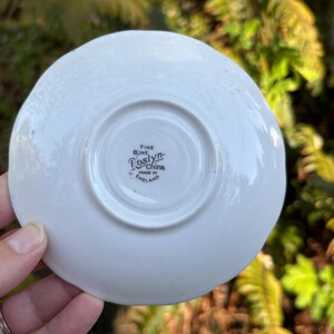 May include: A white, fine bone china saucer with a slightly scalloped edge. The underside features the words "Fine Bone China" and "Toslyn" with "Made in England" below. The saucer is held by a hand.
