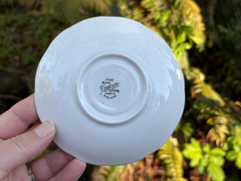 May include: A white, fine bone china saucer with a slightly scalloped edge. The underside features the words "Fine Bone China" and "Toslyn" with "Made in England" below. The saucer is held by a hand.