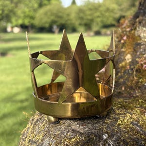 Puede incluir: Un portavelas de metal dorado con forma de corona, con recortes de estrellas. El portavelas tiene una base redonda y tres peque&ntilde;os pies. El fondo es un c&eacute;sped verde borroso y &aacute;rboles.
