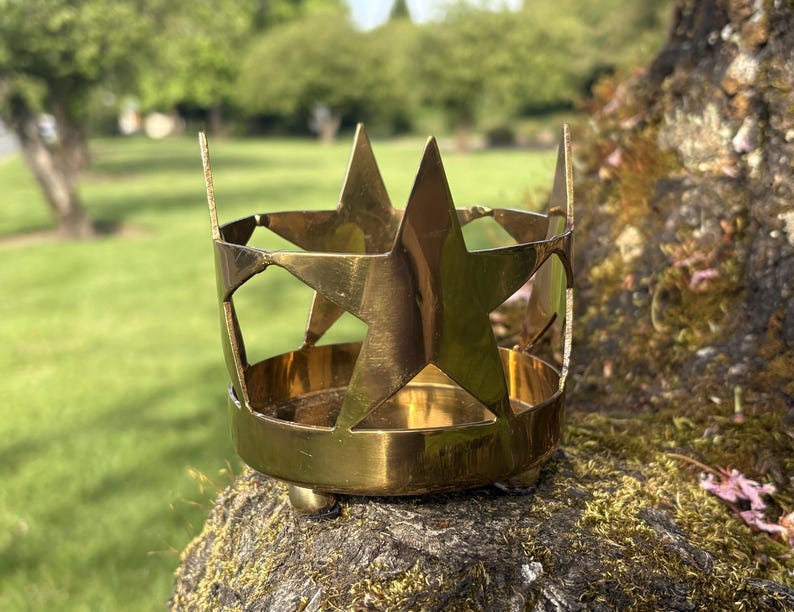 Puede incluir: Un portavelas de metal dorado con forma de corona, con recortes de estrellas. El portavelas tiene una base redonda y tres peque&ntilde;os pies. El fondo es un c&eacute;sped verde borroso y &aacute;rboles.