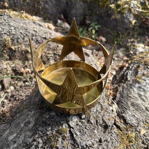 Puede incluir: Un portavelas de color lat&oacute;n con un dise&ntilde;o en forma de estrella. El portavelas tiene una base circular y una estructura en forma de corona con recortes de estrella. Se coloca sobre una superficie natural texturizada.