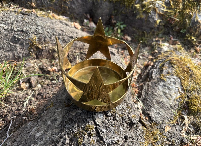 Puede incluir: Un portavelas de color lat&oacute;n con un dise&ntilde;o en forma de estrella. El portavelas tiene una base circular y una estructura en forma de corona con recortes de estrella. Se coloca sobre una superficie natural texturizada.