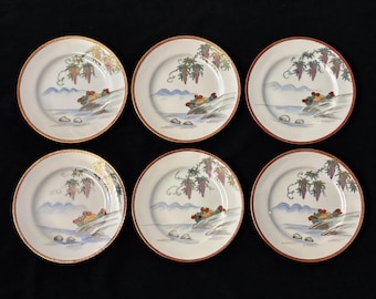 May include: Six white ceramic plates with a decorative design. Each plate features a landscape scene with grapes, a river, and mountains. The plates have a gold rim and a red border.