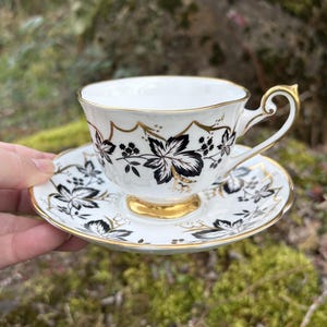 May include: White teacup and saucer set with black floral design and gold trim. The teacup has a curved handle and sits on a matching saucer. The set is held against a blurred green background.