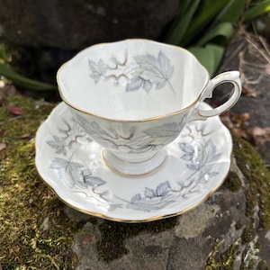 May include: White teacup and saucer set with a gray leaf pattern and gold trim. The teacup has a delicate handle and sits on a matching saucer. The set is made of porcelain and is designed for tea service.