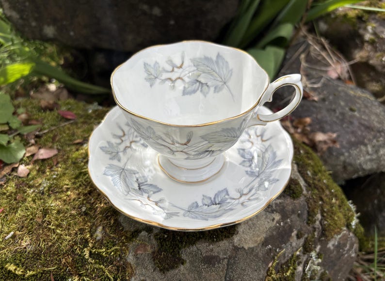 May include: White teacup and saucer set with a gray leaf pattern and gold trim. The teacup has a delicate handle and sits on a matching saucer. The set is made of porcelain and is designed for tea service.